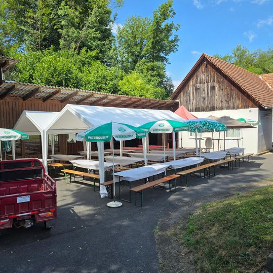 Im Freien gelegener Essbereich mit mehreren Zelten und Bänken, vor einem rustikalen Gebäude. Bäume und ein roter Pickup-Truck sind im Bild.