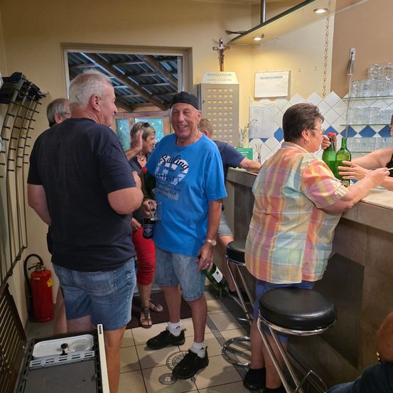 Eine Gruppe von Menschen in einer Kneipe, ein Mann in einem blauen T-Shirt hält ein Glas, eine Frau in einem karierten Shirt hält eine Flasche.