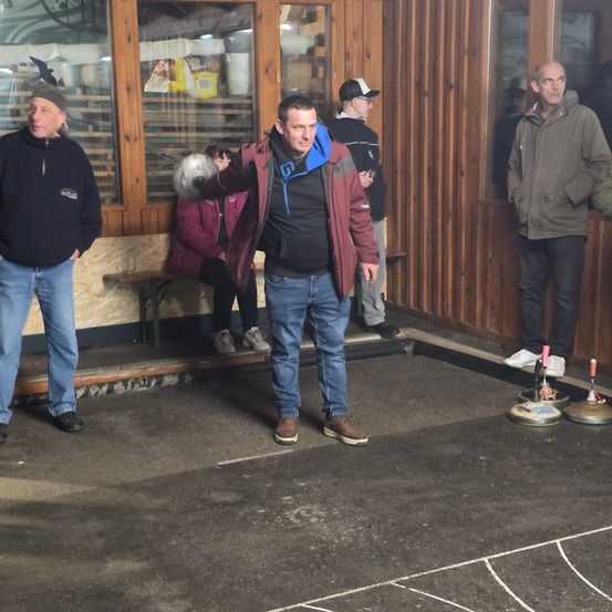 Eine Gruppe von Männern steht herum und beobachtet einen Mann, der auf einer Eisbahn einen Curling-Stein wirft.