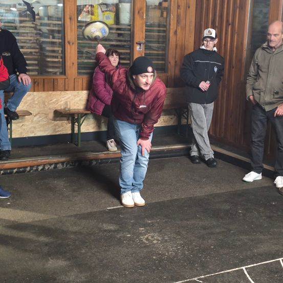 Ein Mann in einer kastanienbraunen Jacke bereitet sich darauf vor, einen Frisbee in einer Außenanlage mit einer Holzwand zu werfen, während andere zusehen.