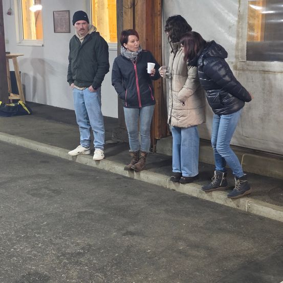 Eine Gruppe von Menschen, darunter ein Mann und drei Frauen, stehen vor einem Gebäude. Eine Frau hält einen Becher und eine andere schaut hinein.