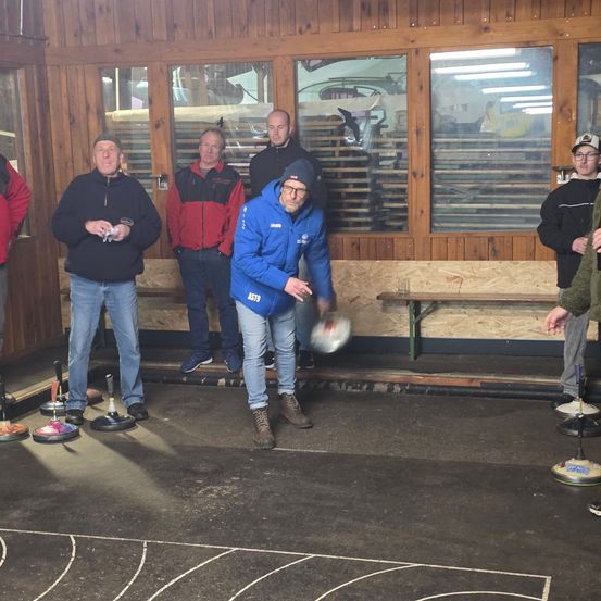 Eine Gruppe von Männern spielt eine Runde Curling in einer hölzernen Halle. Sie tragen Winterkleidung.