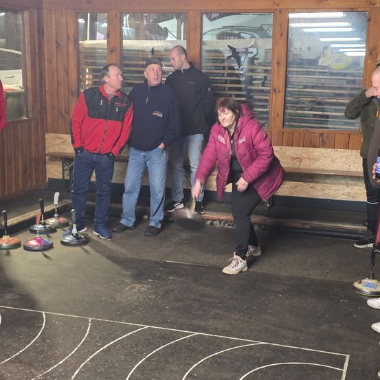 Mehrere Menschen stehen in einem holzverkleideten Raum und beobachten, wie eine Frau einen Curling-Stein wirft. Der Raum hat Bänke und Curling-Ausrüstung.