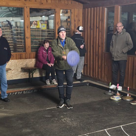 Ein Mann spielt in einem Gebäude eine Partie Curling, während andere zuschauen. Er trägt eine Mütze und eine grüne Jacke.