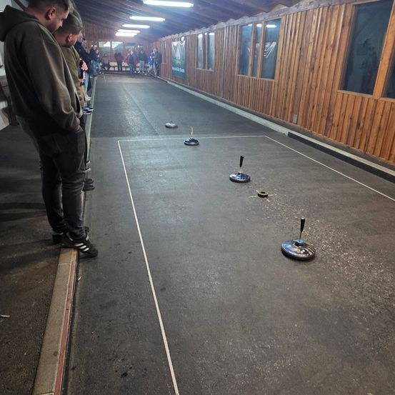 Eine Gruppe von Menschen steht in der Nähe einer Bowlingbahn und beobachtet, wie ein Spiel gespielt wird. Der Platz ist aus Beton und hat Holzwände.