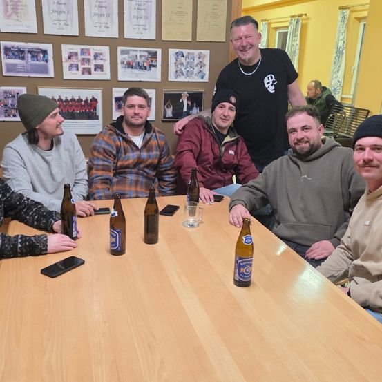 Sechs Männer sitzen um einen Tisch herum mit Bierflaschen und Telefonen. Dahinter eine Wand mit Fotos und einem stehenden, lächelnden Mann.