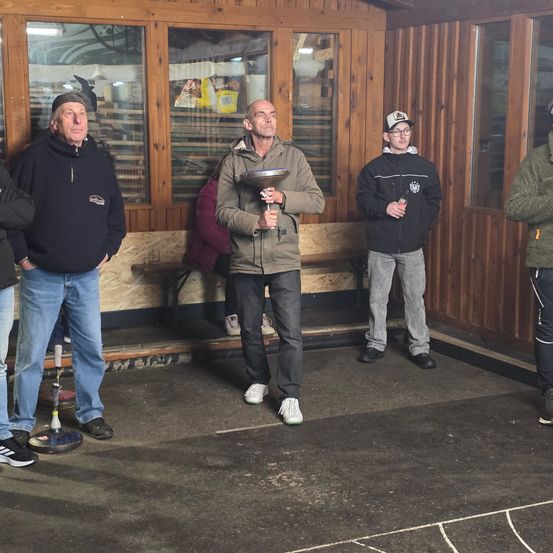 Fünf Männer stehen in einem hölzernen Raum. Der Mann in der Mitte hält eine Trophäe und lächelt. Ein anderer Mann im Hintergrund trägt einen Hut und eine Brille. Sie tragen alle Jacken und Sneakers.