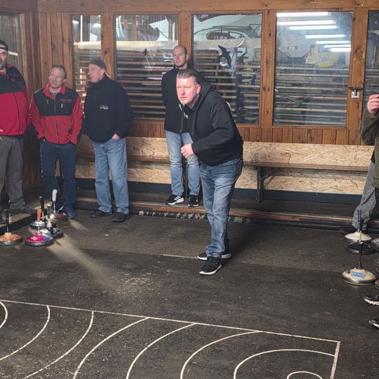 Mehrere Männer in Jacken und Turnschuhen befinden sich in einem Raum mit Holzwänden und Glastüren. Ein Mann wirft einen Curlingstein, während andere zuschauen. Der Boden hat einen Curlingplatz mit einer Kreidelinie.