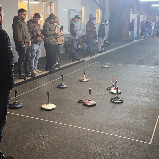 Mehrere Männer warten an den Seitenlinien eines Spielfelds und beobachten ein Spiel. Das Spielfeld hat weiße Linien und mehrere Ringe. Einige Männer halten Flaschen.