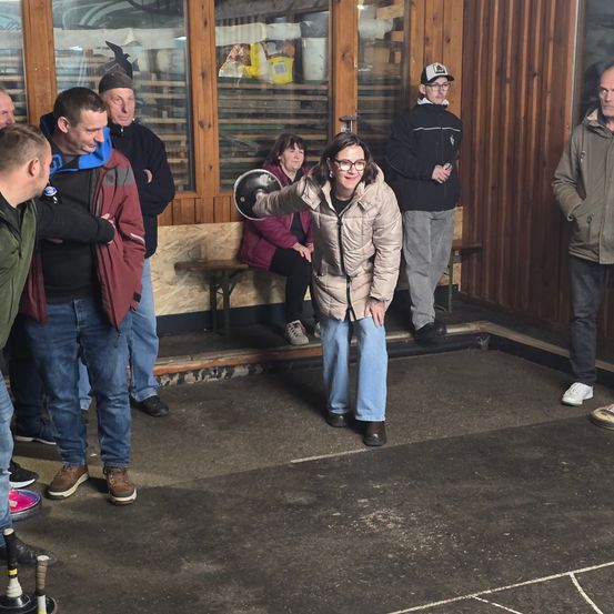 Eine Gruppe von Menschen versammelt sich auf einem Betonbereich, möglicherweise für ein Spiel. Eine Frau mit Brille und einem Ball bereitet sich darauf vor, ihn zu werfen. Einige Leute beobachten sie, einschließlich eines Mannes mit Baseball-Cap. Hinter ihnen befindet sich eine Holzwand mit einer Glastür.