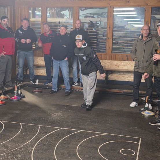 Eine Gruppe von Männern steht in einem Raum und spielt eine Partie Curling. Der Boden hat einen Curling-Platz darauf gezeichnet. Sie tragen alle Jacken und Hosen.