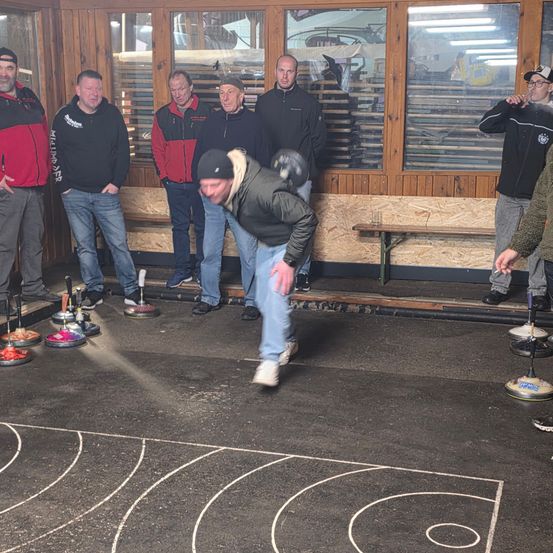Eine Gruppe von Männern spielt Curling auf einer Indoor-Bahn. Ein Mann beugt sich vor, wahrscheinlich um einen Stein zu werfen.