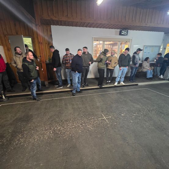 Mehrere Menschen stehen in einer Reihe in einem Raum mit Holzwänden. Einige von ihnen halten Flaschen. Der Raum hat eine weiße Wand mit Fenstern.