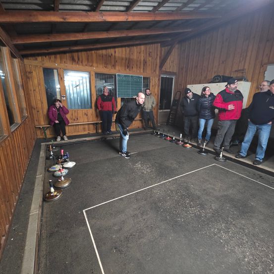 Eine Gruppe von Menschen spielt Curling in einem hölzernen Raum mit schwarzem Boden. Ein Mann wirft einen Curling-Stein.