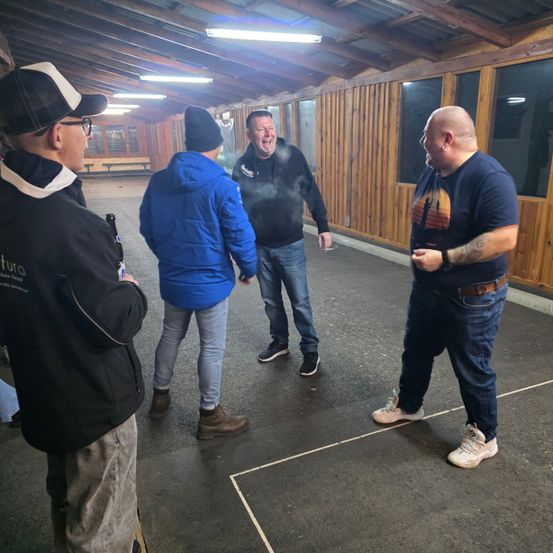 Fünf Männer stehen in einem Raum mit Holzwänden und einer Metalldecke. Ein Mann hält eine Flasche. Ein anderer lacht und bläst Rauch. Der dritte trägt einen Gürtel. Der vierte trägt eine Kappe. Der fünfte trägt eine blaue Jacke.