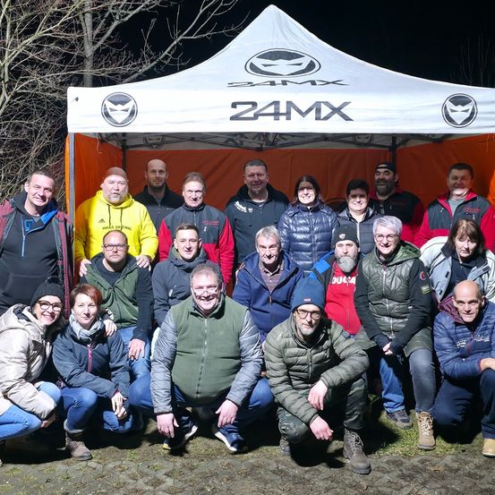 Eine Gruppe von Menschen posiert für ein Foto unter einem weißen Zelt mit der Aufschrift '24MX'. Die Menschen tragen Winterkleidung. Einige von ihnen lächeln, während andere geradeaus schauen. Das Gebiet ist von Gras umgeben.
