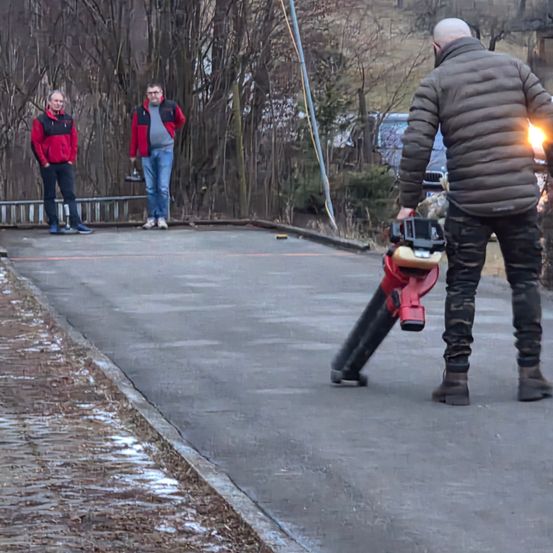 Drei Männer sind draußen, einer hält ein Werkzeug, ein anderer benutzt einen Laubbläser und der dritte steht im Hintergrund.