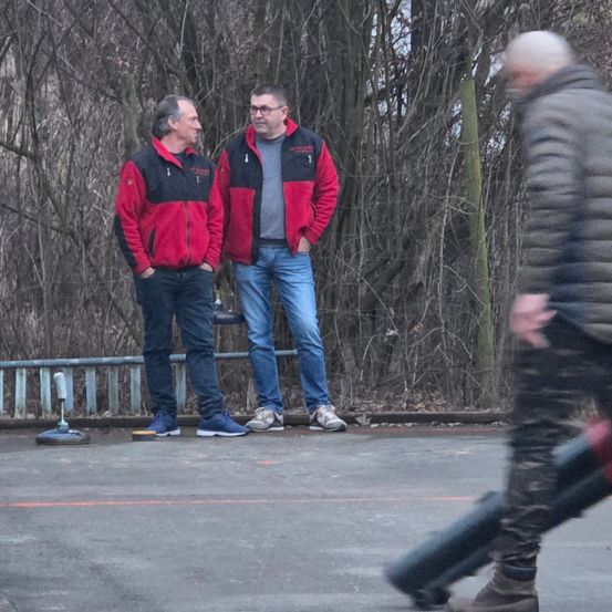 Zwei Männer in roten Jacken stehen in der Nähe eines Zauns und einer Brüstung. Sie schauen etwas vor sich an. Ein Mann in Tarnhose geht auf der Straße.