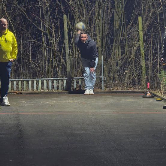 Ein Mann wirft einen Ball während eines Boule-Spiels in der Dunkelheit, während ein anderer Mann zusieht.