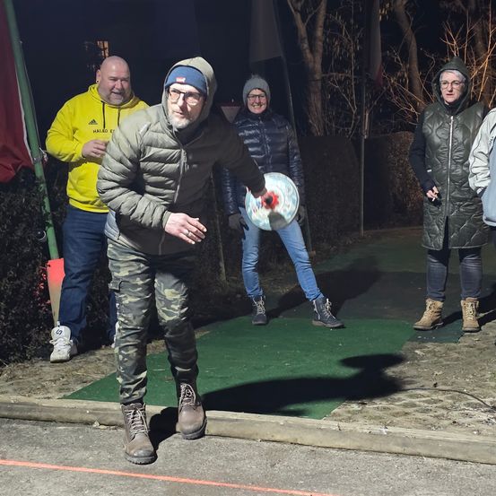 Ein Mann wirft nachts einen Frisbee. Er trägt ein Camouflage-Outfit und eine graue Kapuzenjacke. Ein weiterer Mann in einem gelben Hoodie beobachtet ihn. Eine Frau in einer schwarzen Jacke steht hinter ihm. Eine Flagge ist links. Bäume und Büsche sind im Hintergrund.