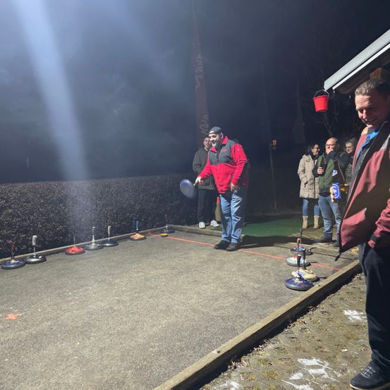 Ein Mann in einem roten Jacken wirft einen Ball auf einem Platz bei Nacht, mit Zuschauern und einem roten Eimer in der Nähe.