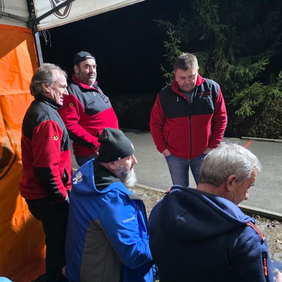 Fünf Männer in roten und blauen Jacken stehen draußen in der Nähe eines Zeltes. Ein Mann trägt eine Mütze, und sie alle schauen in die gleiche Richtung.