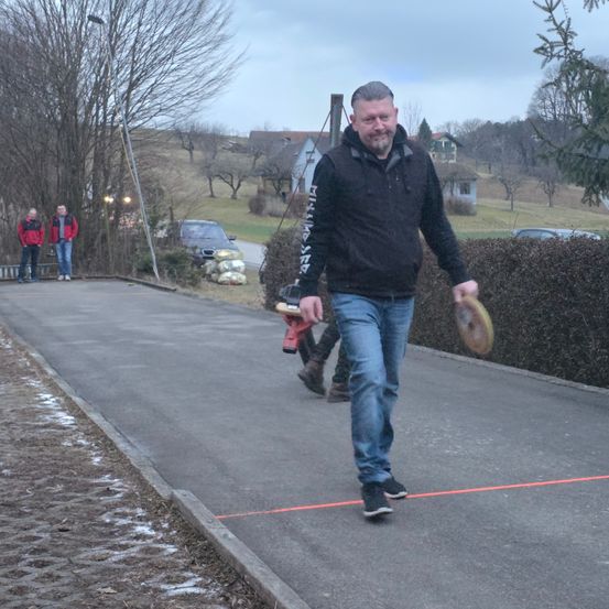 Ein Mann in einer schwarzen Jacke geht auf einem gepflasterten Weg und hält einen Frisbee. Hinter ihm räumt eine Person mit einem roten Laubbläser Blätter auf. Zwei Personen in roten Jacken stehen neben einem Baum.