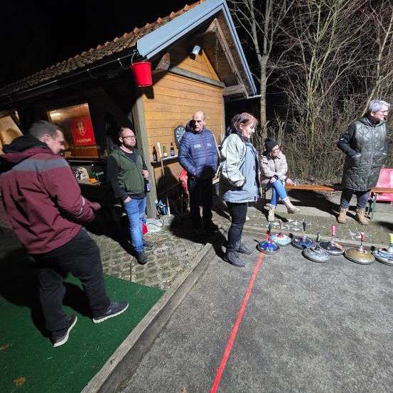 Eine Gruppe von Menschen ist nachts vor einem Gebäude versammelt. Einige stehen, andere sitzen auf einer Bank. Sie scheinen eine Partie Curling zu spielen.