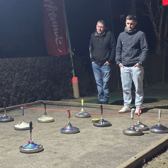 Zwei Männer stehen auf einem Betonfeld mit Curling-Ausrüstung vor ihnen, einem Banner dahinter und einem blattlosen Baum.