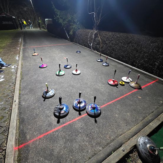 Eine nächtliche Szene zeigt eine Reihe von farbenfrohen Ringen, die auf einem Betonweg angeordnet sind, mit roten Markierungen. Menschen stehen an den Rändern.