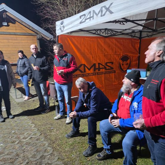 Eine Gruppe von Menschen steht und sitzt vor einem Zelt mit einer orangefarbenen Fahne. Sie sind in Winterkleidung gekleidet. Ein Mann spricht und hält ein Getränk in der Hand.