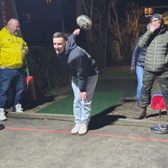 Ein Mann in einer blauen Kapuzenjacke wirft nachts im Freien einen Frisbee, beobachtet von drei anderen. Die Gruppe steht auf einer Straße mit einer roten Linie und einem grünen Grasfleck.