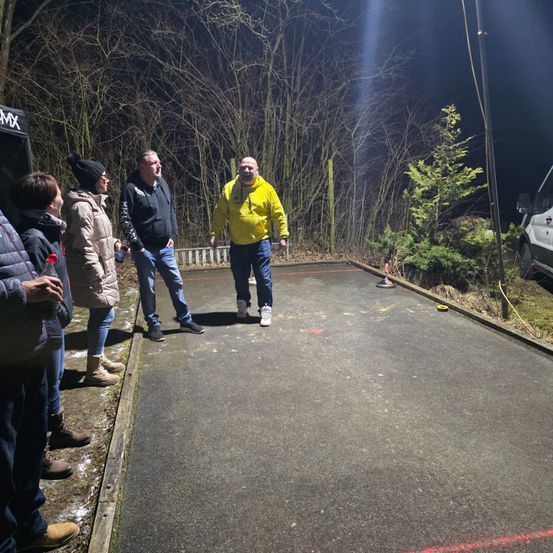 Eine Gruppe von Menschen steht in einem schwach beleuchteten Bereich, ein Mann läuft auf einem Betonweg, Bäume und ein weißer Van im Hintergrund.