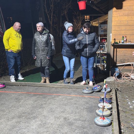 Vier Personen stehen draußen auf einem gepflasterten Bereich mit einer Reihe von Curling-Steinen, ein Mann in einer gelben Jacke und drei Frauen in Wintermänteln.