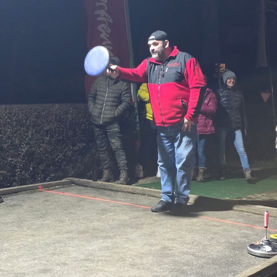 Ein Mann in einem rot-schwarzen Jacken wirft einen blauen Frisbee auf einem Betonbereich, während andere zuschauen.