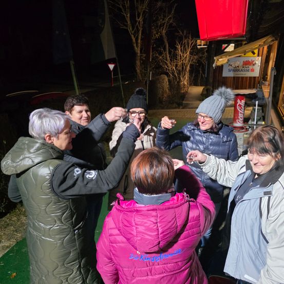 Eine Gruppe von Frauen steht bei Nacht im Freien, lächelt und hebt ihre Gläser. Sie sind in Winterjacken gekleidet und einige tragen Mützen. Im Hintergrund steht ein Schild mit der Aufschrift 'SPARKASSE'.