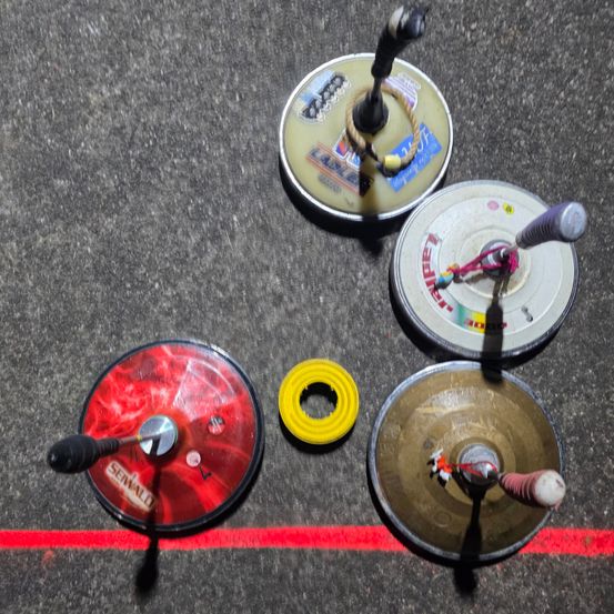 Fünf runde Objekte mit verschiedenen Designs und Farben liegen auf einer Betonoberfläche. Vier haben Griffe und eines ist gelb. Eine rote Linie verläuft durch die Mitte.