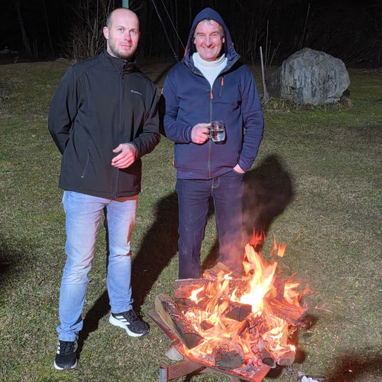 Zwei Männer stehen neben einem Lagerfeuer in der Nacht. Einer hält ein Getränk. Ein großer Fels ist im Hintergrund.