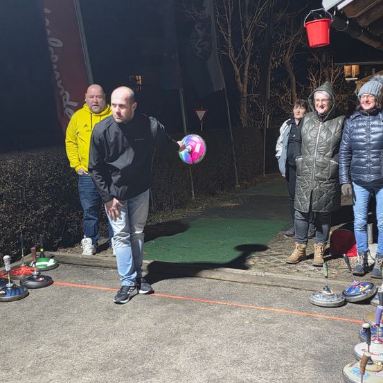 Eine Gruppe von Menschen spielt draußen bei Nacht eine Partie Curling. Ein Mann bereitet sich darauf vor, einen Ball zu werfen.