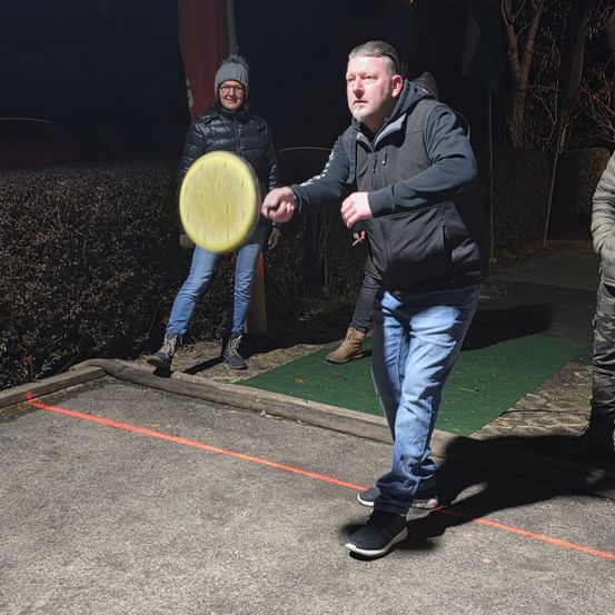 Ein Mann wirft nachts in einem Park einen Frisbee, während eine Frau im Hintergrund zuschaut.