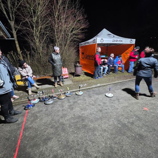 Bei Nacht sind Menschen um ein Zelt mit 24MX-Marke versammelt. Einige sitzen auf einer Bank, andere stehen. Ein paar Objekte, die Rollern ähneln, liegen auf dem Boden.