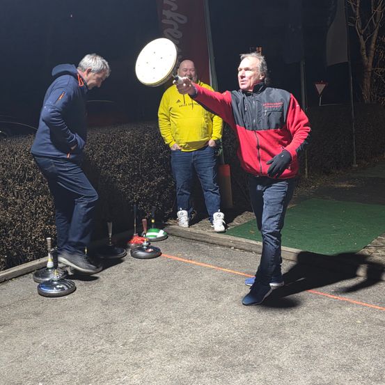 Drei Männer stehen bei Nacht draußen, einer in einem rot-schwarzen Jacken wirft eine Scheibe, ein anderer beugt sich und ein dritter beobachtet. Sie stehen auf einem Betonbereich mit Hecken und einer grünen Matte.