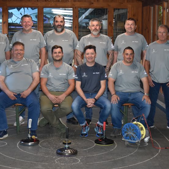 A group of men in gray shirts and jeans are posing for a photo inside a wooden room with a circle on the floor.