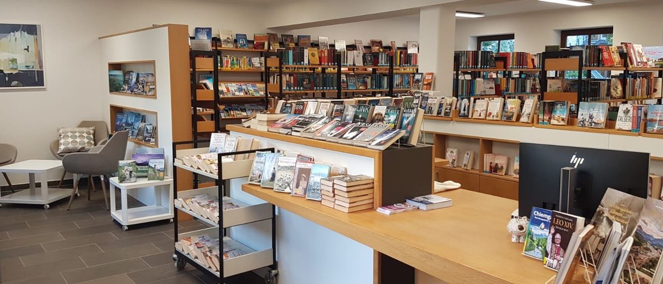 Eine Buchhandlung mit Bücherregalen aus Holz. Ein Wagen mit Büchern auf Rädern und ein Tisch mit Büchern. Es gibt Deckenleuchten.