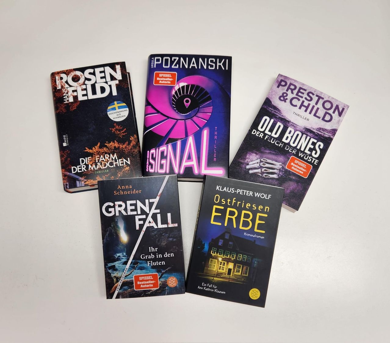 Fünf Bücher sind auf einer weißen Oberfläche ausgelegt. Von links nach rechts sind die Titel: Rosenfeld, Signal, Grenzfall, Old Bones und Ostfriesen Erbe.