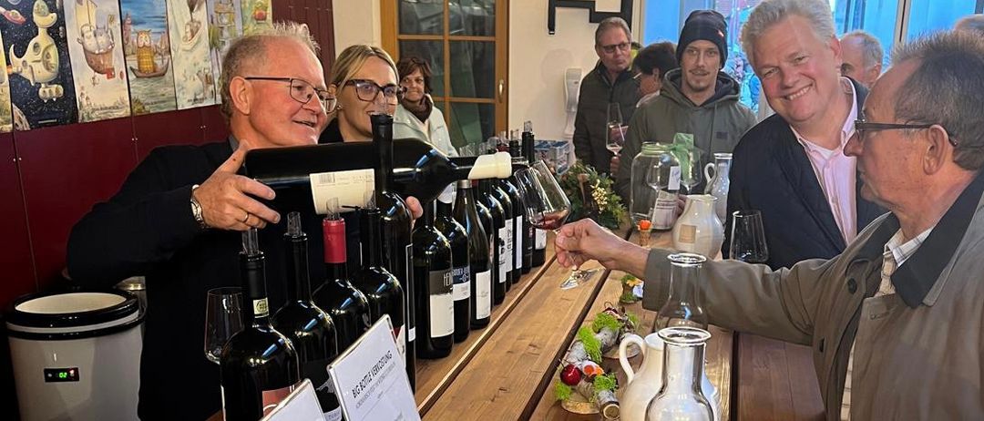 Eine Gruppe von Menschen, darunter zwei Männer, sitzen in einem Restaurant an einem Tisch. Ein Mann schenkt Wein in ein Glas. Sie sind umgeben von Weinflaschen, Weingläsern und anderen Gegenständen. Der Tisch hat Schilder mit der Aufschrift 'Big Bottle Verkostung'. An der Wand hängen auch Gemälde.