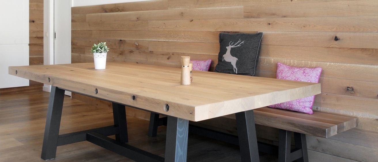 Ein Esstisch mit einer Holzbank und einer Holzwand. Ein Topf mit Pflanzen steht auf dem Tisch. Ein Kissen mit Hirschdesign liegt auf der Bank.