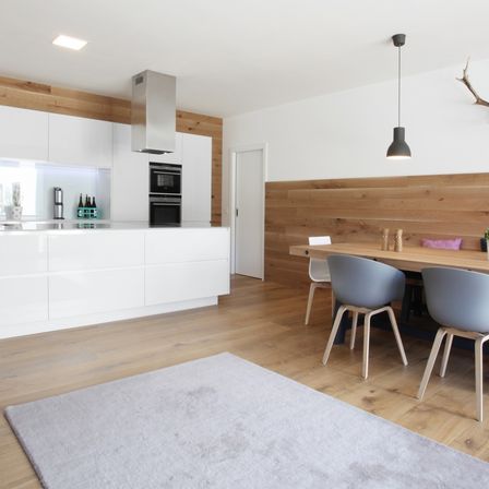 Moderne Küche mit weißen Schränken, Holzboden und einem Essbereich mit Stühlen und Tisch. Eine Lampe hängt von der Decke.