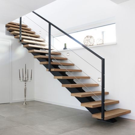 Eine moderne Treppe mit Holzstufen und schwarzen Metallhandläufen. Der Raum ist hell mit weißen Wänden und einem Fenster mit Blick auf eine Pflanze.