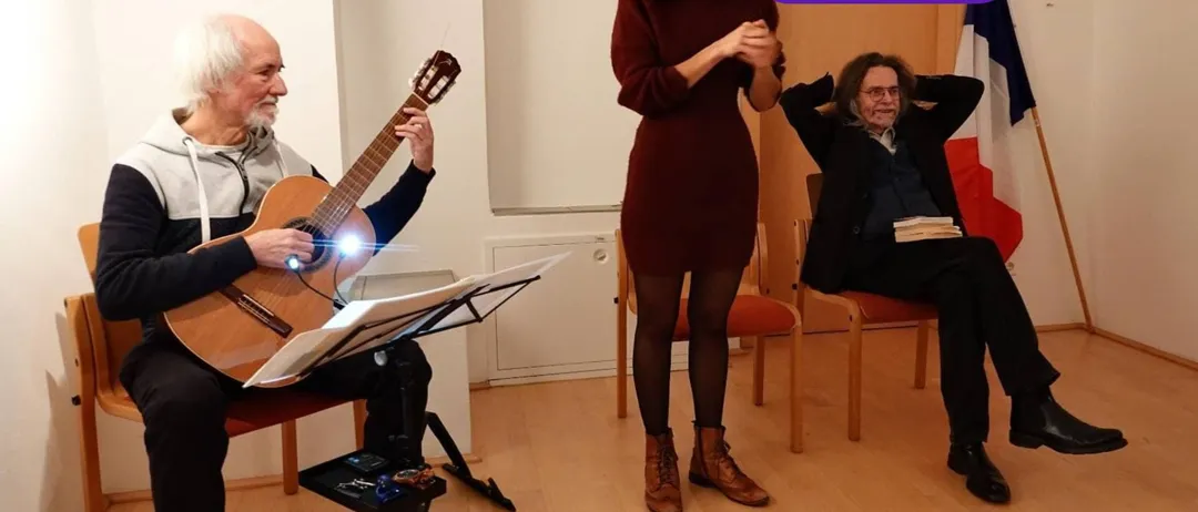Ein älterer Mann spielt Gitarre, während eine Frau vor ihm singt. Ein anderer Mann sitzt auf einem Stuhl hinter ihnen.
